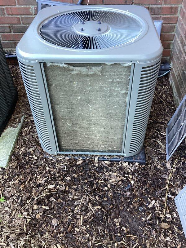 Completed annual preventative maintenance on residential split system air conditioning unit. Inspected condenser unit exterior, confirmed proper fan guard assembly and structural integrity, verified unit positioning and clearances around equipment. Noted heavy dirt and debris accumulation on front condenser coil requiring cleaning attention. Documented unit condition through photographic records showing outdoor installation against brick foundation with adequate mulch bed clearance and proper concrete pad placement.
