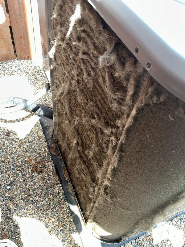 # HVAC Maintenance - ACM Completed

Performed comprehensive air conditioning maintenance on outdoor condenser unit. Upon arrival, discovered extremely heavy accumulation of lint, dust, and fibrous debris coating the exterior condenser coils and side panels. The buildup was significantly restricting airflow and indicated extended period of maintenance neglect, with thick matted layers covering nearly all exposed coil surfaces.

Thoroughly cleaned condenser coils using appropriate methods to remove the dense accumulation of debris from all accessible surfaces. Cleared obstructions from the unit's metal fins and exterior panels to restore proper airflow capacity. Inspected unit mounting, concrete pad, and surrounding gravel base for stability and drainage. Verified PVC condensate line routing and condition. System operational following cleaning and maintenance procedures.