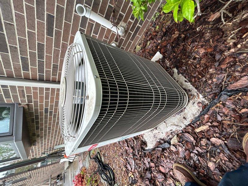 Completed annual cooling maintenance on residential outdoor condenser unit. Inspected cylindrical condenser positioned on concrete pad adjacent to brick building exterior. Verified proper operation of top-mounted fan assembly protected by wire mesh grille. Examined condenser coil fins for damage, debris, and blockage—fins found in good condition with adequate airflow clearance. Checked refrigerant lines and electrical conduit routing along building wall for secure mounting and proper insulation. Inspected PVC condensate drain line for proper drainage and confirmed no obstructions. Replaced furnace filters as part of routine maintenance protocol. Cleared surrounding mulch bed area of excessive organic debris to maintain adequate airflow around unit. Verified unit operational parameters within manufacturer specifications. System tested and confirmed cooling cycle functioning properly with no abnormalities detected.