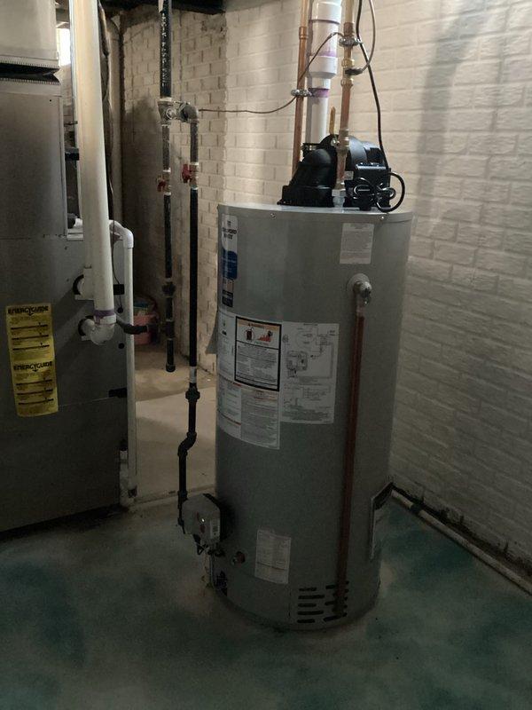 Successfully installed a 50-gallon tanked water heater in basement utility room. Removed existing unit and positioned new water heater on concrete floor, ensuring proper clearances and level placement. Connected hot and cold water supply lines using copper piping with appropriate shut-off valves and dielectric unions. Installed recirculation pump assembly on top of unit with associated black pump mechanism and PVC discharge piping routed vertically through ceiling joists. Made all necessary electrical connections to power the unit and pump system. Installed temperature and pressure relief valve with proper discharge piping termination. Tested all water connections for leaks, verified proper venting configuration, and ensured all plumbing code compliance requirements were met. Confirmed unit operational functionality including heating cycle, thermostat response, and recirculation pump operation. All manufacturer labels and specifications remain visible on front of tank for future refere
