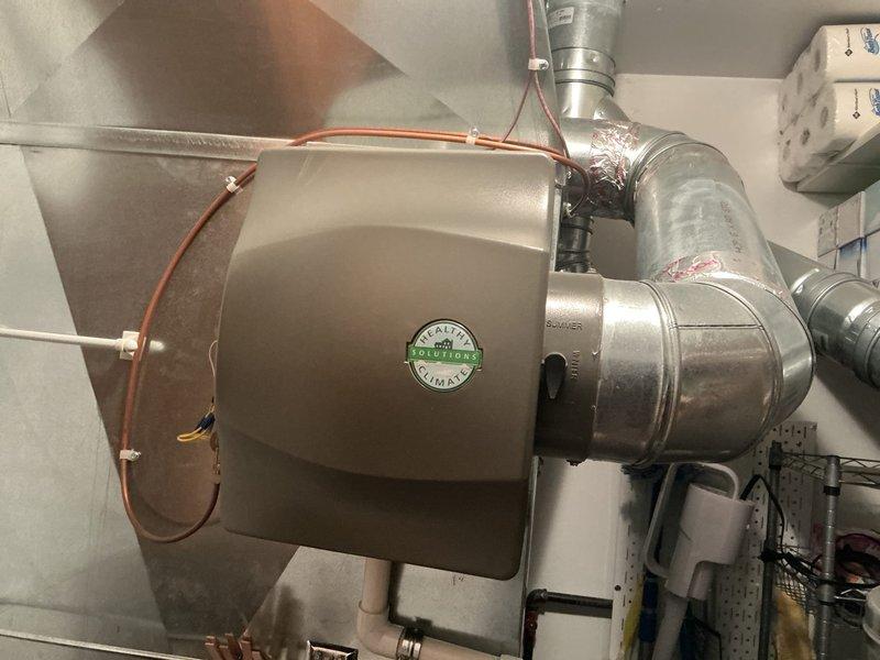 Completed annual HVAC maintenance inspection on residential air conditioning system. Inspected whole-house humidifier unit mounted in mechanical room, verifying proper installation, ductwork connections, and water supply lines. Documented equipment condition with photographic evidence showing unit mounted to ductwork with copper water lines intact. Inspected return air grille and noted accumulated dust, debris, and cobweb material on horizontal slats requiring cleaning attention. All major components operational with no immediate safety concerns identified during visual inspection.