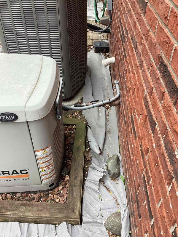 Responded to service call for generator and air conditioning unit sinking into ground. Upon arrival, inspected 17kW residential standby generator and adjacent AC condensing unit positioned against brick wall foundation. Found both units settling unevenly into gravel base, with weathered landscape timber border deteriorating and providing inadequate support. Assessed structural stability of installation, noting compromised positioning affecting proper drainage and potential stress on refrigerant line connections and electrical conduit. Documented current conditions including copper refrigerant lines with foam insulation, PVC piping, and mounting configuration. Photographed equipment placement, ground settling patterns, and existing infrastructure to establish baseline for corrective action. Recommended installation of concrete pad or reinforced gravel base with proper compaction to prevent further settling and ensure manufacturer specifications for level mounting are maintained.