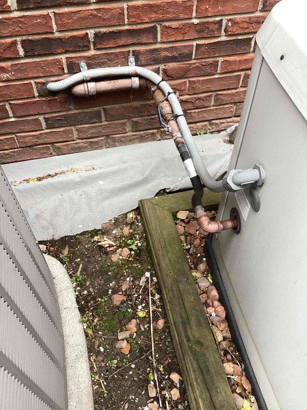 Responded to service call for generator and air conditioning unit sinking into ground. Upon arrival, inspected 17kW residential standby generator and adjacent AC condensing unit positioned against brick wall foundation. Found both units settling unevenly into gravel base, with weathered landscape timber border deteriorating and providing inadequate support. Assessed structural stability of installation, noting compromised positioning affecting proper drainage and potential stress on refrigerant line connections and electrical conduit. Documented current conditions including copper refrigerant lines with foam insulation, PVC piping, and mounting configuration. Photographed equipment placement, ground settling patterns, and existing infrastructure to establish baseline for corrective action. Recommended installation of concrete pad or reinforced gravel base with proper compaction to prevent further settling and ensure manufacturer specifications for level mounting are maintained.