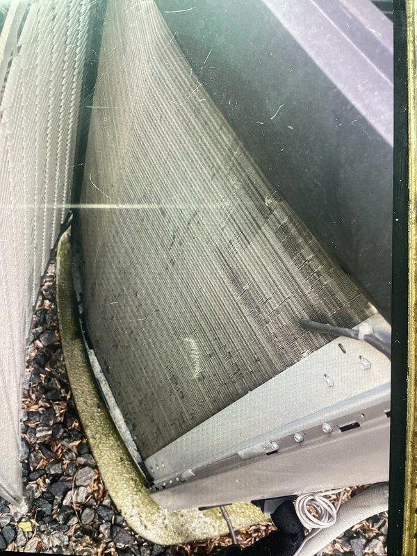 # HVAC Maintenance - ACM Completed

Performed annual cooling maintenance on outdoor air conditioning condenser unit. Conducted comprehensive visual inspection of equipment, noting minor dust and debris accumulation on condenser coils and interior surfaces. Observed leaves and organic debris collected around unit base and concrete pad perimeter. Inspected evaporator/condenser coil assembly, confirming curved fin structure intact with characteristic ribbed pattern present. Verified proper mounting on concrete pad with all fasteners secure. Examined electrical connections and wiring, noting white electrical cord properly positioned beneath unit. Confirmed warning labels and safety placards remain legible and properly affixed to metal housing. Documented equipment condition through photographic records showing top-down view of coil assembly and exterior side view of unit. Unit operational with gray metal casing and horizontal fin coils visible through ventilation openings in serviceable co