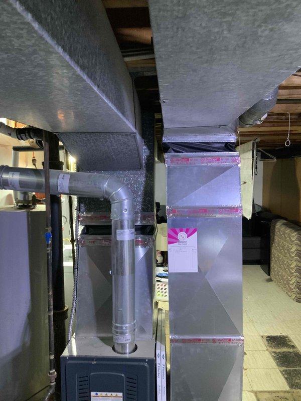 Conducted site assessment for air conditioning system installation at residential property with existing furnace and ductwork infrastructure but no previous cooling system. Evaluated basement mechanical room containing dark-colored furnace unit with connected vertical ductwork, rectangular air handler, and foil-faced insulated duct runs throughout ceiling space. Inspected exterior location near glass block foundation windows for potential condenser unit placement. Provided comprehensive estimate for complete AC system installation utilizing existing ductwork distribution network.