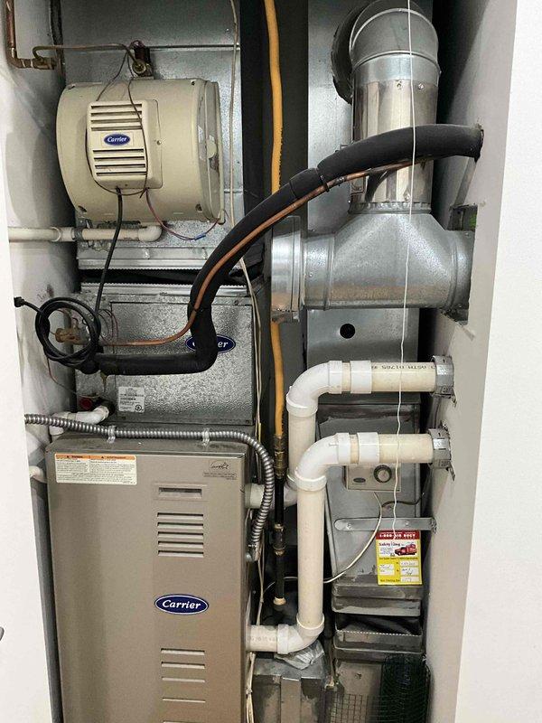 Provided comprehensive estimate for replacement of 20+ year old Carrier furnace and air conditioning system currently located on main floor in utility closet or mechanical room. Existing natural gas furnace remains operational but has reached end of expected service life. Current installation includes Carrier furnace with integrated Aprilaire humidifier, extensive ductwork configuration with galvanized metal elbows, stainless steel venting, white PVC condensate drain lines, and complete electrical connections. Documented existing system layout including vertical furnace placement, wall-mounted humidification equipment, draft inducer assembly, flexible ducting, and all associated piping for proper sizing and replacement specifications.