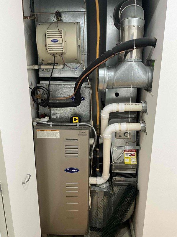 Provided comprehensive estimate for replacement of 20+ year old Carrier furnace and air conditioning system currently located on main floor in utility closet or mechanical room. Existing natural gas furnace remains operational but has reached end of expected service life. Current installation includes Carrier furnace with integrated Aprilaire humidifier, extensive ductwork configuration with galvanized metal elbows, stainless steel venting, white PVC condensate drain lines, and complete electrical connections. Documented existing system layout including vertical furnace placement, wall-mounted humidification equipment, draft inducer assembly, flexible ducting, and all associated piping for proper sizing and replacement specifications.