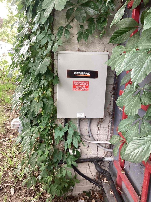 Responded to service call for inoperative Generac generator system approximately 2 years old and covered under 10-year manufacturer warranty. Upon arrival, located exterior-mounted Generac automatic transfer switch on building exterior wall with multiple conduits and connections. Performed visual inspection of transfer switch enclosure and noted significant vegetation overgrowth around unit requiring clearance for proper access and ventilation. Documented current installation conditions with photographs showing transfer switch mounting, conduit routing, and surrounding environment. Verified warranty coverage status and reviewed service eligibility with customer regarding dispatch fees and warranty terms. Assessed transfer switch operation and electrical connections to diagnose cause of generator failure to start or transfer load during power outage conditions.