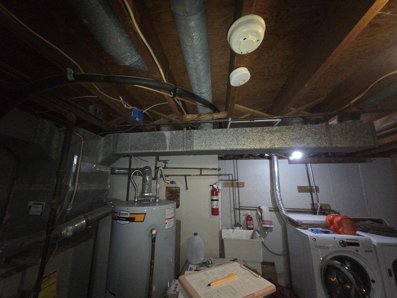 Conducted comprehensive assessment for HVAC system replacement in basement utility area. Evaluated existing ductwork components and connections, noting metal cylindrical ducts with ribbed texture and round connectors requiring replacement. Documented current system configuration including proximity to water heater and other mechanical systems, with attention to ceiling clearance and existing utility connections for proper sizing of replacement unit.
