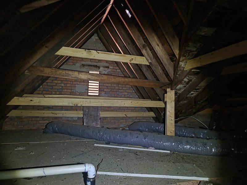 Conducted comprehensive attic inspection to assess existing air conditioning system and provide replacement estimate. Evaluated attic-mounted HVAC equipment, existing ductwork configuration, structural access points, and insulation conditions. Documented current system layout including flexible duct routing, PVC condensate drainage, ventilation pathways, and available clearances for equipment replacement. Photographed attic space showing wooden framing, existing black flexible ductwork, insulation materials, and brick chimney structure to support replacement recommendations and equipment sizing calculations.