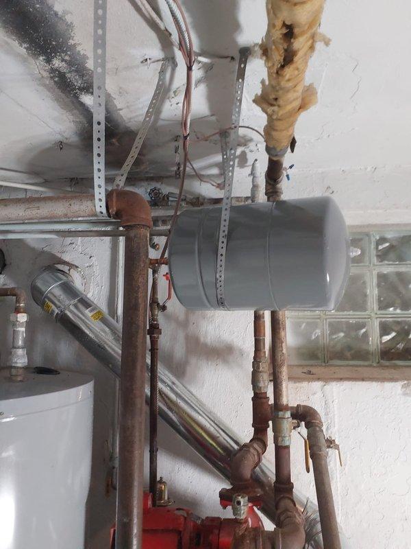 Installed new expansion tank and additional copper piping to commercial heating system in utility room. Mounted gray cylindrical expansion tank horizontally to wall using metal brackets and strapping, connecting to existing copper pipe network with appropriate fittings and red-handled isolation valves. Completed all necessary piping connections to integrate expansion tank with heating system components, ensuring proper pressure relief capability for the hydronic system.