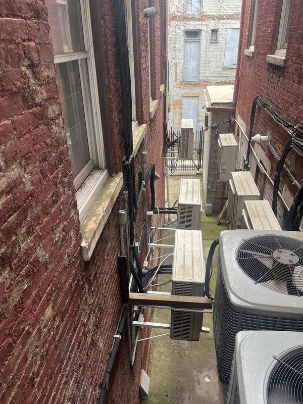Conducted on-site estimate for commercial HVAC replacement involving three ductless mini-split systems and one air conditioning unit. Assessed existing equipment located in exterior alleyway courtyard area, noting multiple aging AC units positioned between brick buildings with visible weathering and environmental exposure. Evaluated interior space requiring mini-split installation in kitchen/dining area where one wall-mounted unit is currently installed. Documented equipment locations, building access points, and spatial constraints for proposed system installations in multi-unit urban building environment.