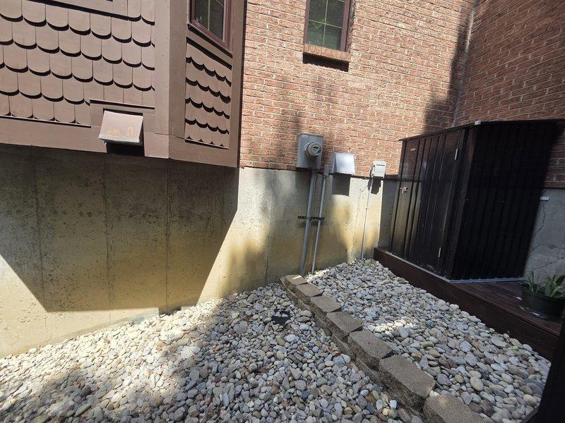 Conducted site evaluation for geothermal heating system replacement. Documented existing hydronic heating infrastructure including hydro separator with loop connections and dual Grundfos circulation pumps mounted on plywood backing, as well as exterior ground loop access points and utility connections on brick exterior with river rock landscaping.