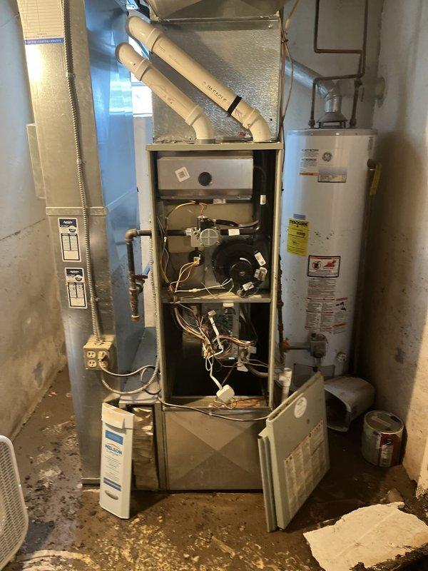 Conducted on-site assessment of flooded HVAC system in residential utility room. Inspected furnace unit with front panel removed, exposing blower motor, internal wiring, and mechanical components affected by water damage. Evaluated condition of beige metal cabinet furnace with PVC exhaust/intake venting, associated ductwork, and electrical connections. Assessed water heater positioned adjacent to heating system for potential impact from flooding incident. Documented extensive water damage to concrete floor area and equipment. Prepared comprehensive replacement estimate for complete HVAC system replacement due to flood damage rendering existing equipment non-serviceable.