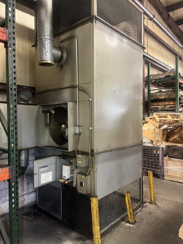 Conducted on-site estimate for commercial furnace replacement at industrial facility. Unit is a ground-mounted packaged heating system serving the factory area, showing significant age and wear with visible dirt accumulation, surface staining, and weathering on exterior metal panels. Assessed existing beige metal cabinet-style unit with ventilation grilles, galvanized ductwork connection, blower assembly, and electrical control panel protected by safety barriers. Documented equipment condition through photographs and evaluated replacement requirements for continued operation of facility heating system.