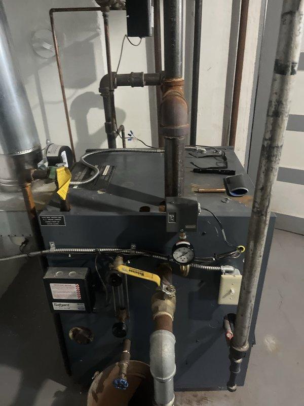 Conducted comprehensive assessment of existing steam boiler system for replacement estimate. Evaluated aging unit with visible corrosion on piping infrastructure, documented current equipment configuration including pressure gauge, valve assemblies, and connection points. Photographed existing installation showing dark blue/gray metal cabinet unit with multiple pipe connections including copper supply lines, PVC drainage, and black iron distribution piping. Assessed overall system condition, noting weathered components and oxidation on metal surfaces throughout the mechanical room installation.
