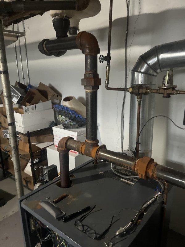 Conducted comprehensive assessment of existing steam boiler system for replacement estimate. Evaluated aging unit with visible corrosion on piping infrastructure, documented current equipment configuration including pressure gauge, valve assemblies, and connection points. Photographed existing installation showing dark blue/gray metal cabinet unit with multiple pipe connections including copper supply lines, PVC drainage, and black iron distribution piping. Assessed overall system condition, noting weathered components and oxidation on metal surfaces throughout the mechanical room installation.