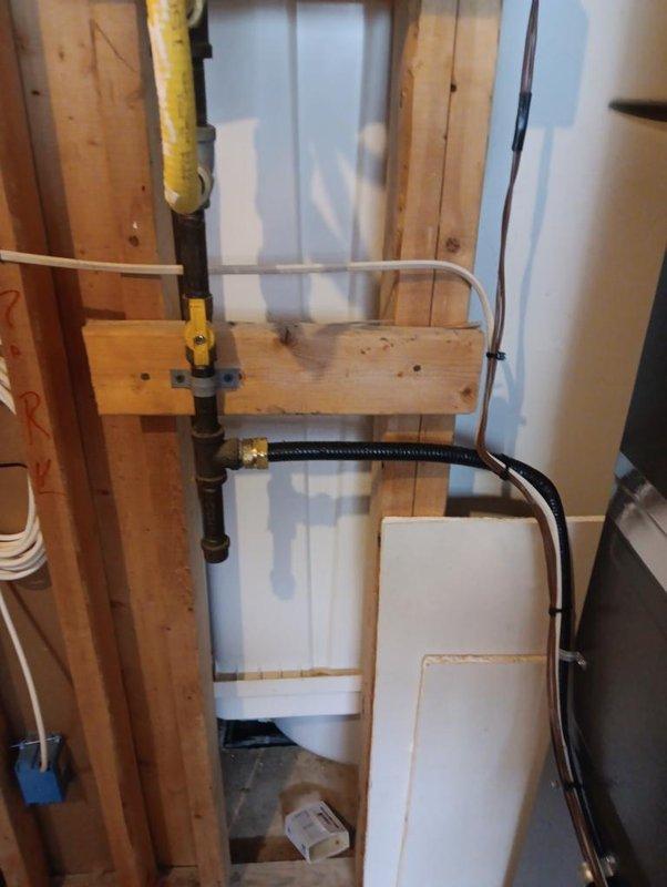 Completed installation of gas line and electrical panel in residential construction area. Installed black metal gas pipe with brass connector and flexible braided high-pressure line for appliance connection, routed yellow flexible conduit through wooden framing, and mounted electrical panel box with circuit breakers on exposed wall studs with proper wire management through insulated cavity.