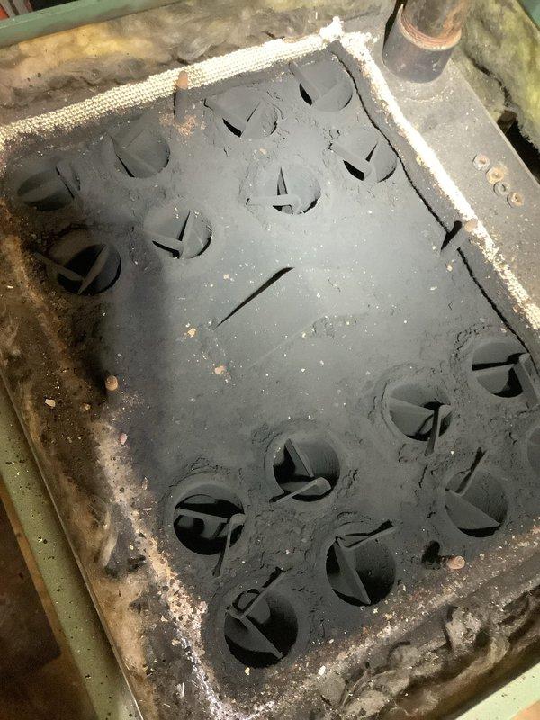 Responded to heating system service call and found the ventilation panel had detached from the oil heating unit and was lying on the floor. Inspected the heat exchanger assembly, which showed significant mineral deposits, scale buildup, and corrosion throughout the component and surrounding frame, indicating extended service life and moisture exposure. Documented the condition of the system with photographs showing the deteriorated ventilation panel with accumulated dust and debris, as well as the heavily scaled burner assembly with corrosion around circular openings and ductwork connections.