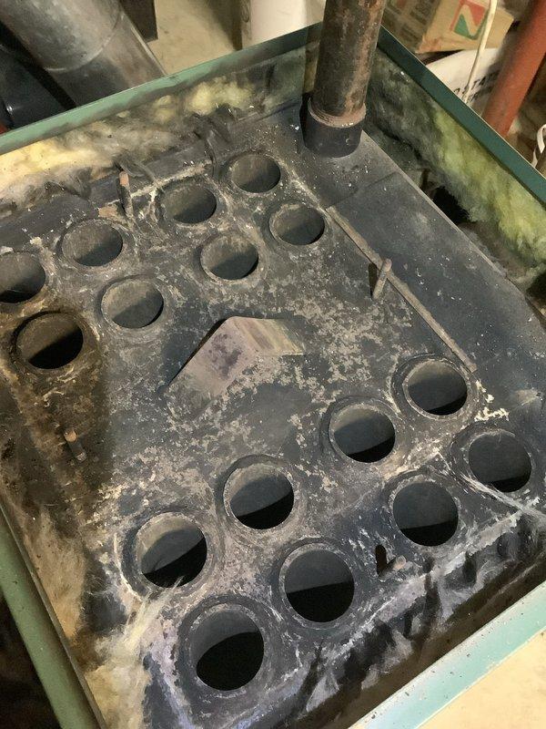 Responded to heating system service call and found the ventilation panel had detached from the oil heating unit and was lying on the floor. Inspected the heat exchanger assembly, which showed significant mineral deposits, scale buildup, and corrosion throughout the component and surrounding frame, indicating extended service life and moisture exposure. Documented the condition of the system with photographs showing the deteriorated ventilation panel with accumulated dust and debris, as well as the heavily scaled burner assembly with corrosion around circular openings and ductwork connections.