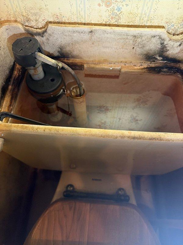 Responded to dispatch for continuously running toilet on ground floor unit. Upon inspection, removed tank cover and found fill valve assembly requiring adjustment. Adjusted standpipe height to correct water level. Tank interior showed extensive rust staining, mineral deposits, and mold growth indicating long-term water exposure and lack of maintenance. Fill valve with red cap and float mechanism were functional but corroded. Black rubber flapper and chain assembly showed signs of deterioration. After adjustment, toilet continued running, indicating additional repair needs beyond initial standpipe adjustment. Tank cover could not be properly reinstalled due to component interference. Documented tank condition showing heavy rust on porcelain surfaces, corroded brass fittings, discolored water, and significant biological growth along tank rim requiring comprehensive cleaning and potential component replacement for complete repair.