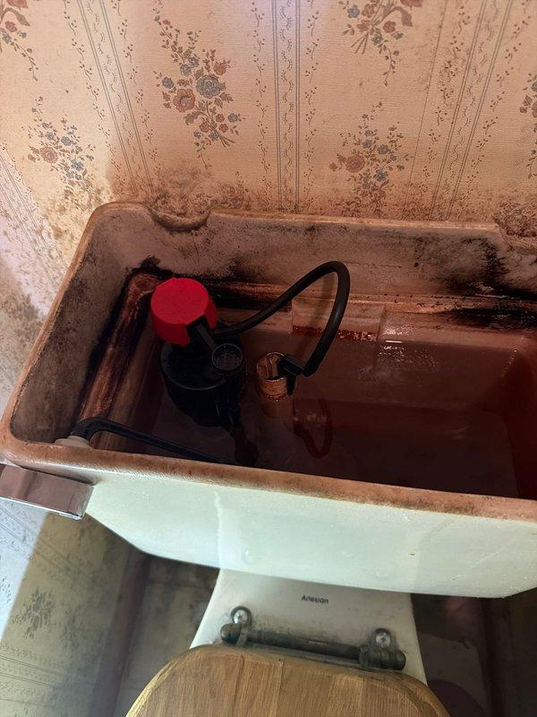 Responded to dispatch for continuously running toilet on ground floor unit. Upon inspection, removed tank cover and found fill valve assembly requiring adjustment. Adjusted standpipe height to correct water level. Tank interior showed extensive rust staining, mineral deposits, and mold growth indicating long-term water exposure and lack of maintenance. Fill valve with red cap and float mechanism were functional but corroded. Black rubber flapper and chain assembly showed signs of deterioration. After adjustment, toilet continued running, indicating additional repair needs beyond initial standpipe adjustment. Tank cover could not be properly reinstalled due to component interference. Documented tank condition showing heavy rust on porcelain surfaces, corroded brass fittings, discolored water, and significant biological growth along tank rim requiring comprehensive cleaning and potential component replacement for complete repair.