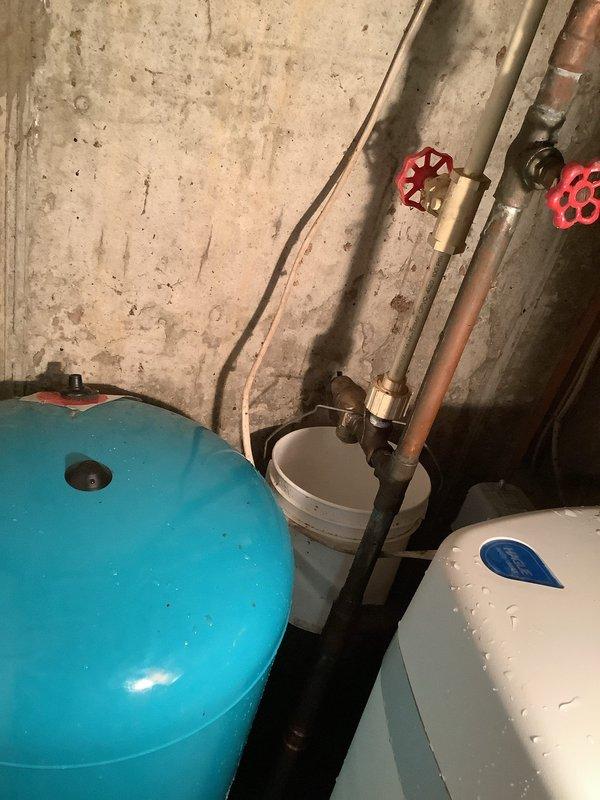 Responded to service call for active drip/leak at temperature and pressure relief valve on well pressure tank system. Upon inspection, confirmed leak at T&P relief valve near well tank installed in 2018. Photographed existing conditions showing turquoise pressure tank, white expansion tank, copper piping with shut-off valves, and pressure gauge assembly. Placed collection bucket beneath leak to prevent water damage. Assessed system components and documented leak source for repair recommendation.