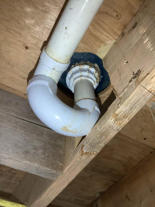 Responded to service call for reported leak near condensate pump and air handler assembly. Upon inspection, located active water leak at P-trap connection in condensate drain line beneath air handler unit. Identified compromised pipe joint and deteriorated rubber boot seal at floor penetration point allowing condensate water to escape. Evaluated drain line configuration and structural framing around affected area to determine proper repair approach. Documented existing conditions with photographs showing P-trap assembly, PVC drain piping, floor joists, and compromised sealing components for service records and future reference.