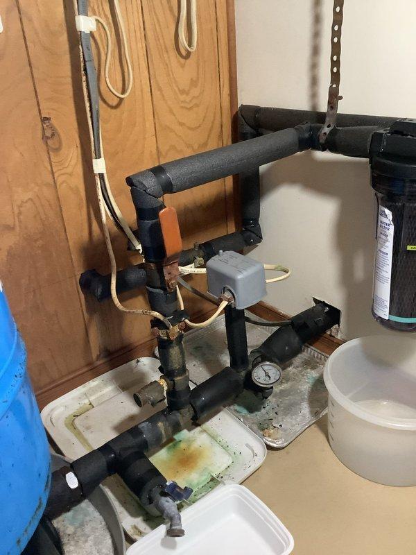 Responded to troubleshoot ongoing water system issues including recurring low water pressure, filter contamination with black sediment, and persistent leaking at the whole house water filter assembly. Conducted comprehensive inspection of the complete water system from well to point-of-entry equipment, examining all plumbing connections, pressure regulation components, and filtration hardware. Documented existing conditions with photographs showing the insulated pipe network, pressure gauge, filter housing with visible leak collection pans positioned beneath the assembly, and associated water treatment components. Identified the whole house filter as the primary source of leaking, which had progressed from an initial leak that temporarily stopped after filter replacement to a current slow drip condition. Noted significant black sediment accumulation requiring multiple filter changes within a short timeframe, indicating potential issues with well water quality or system contamination. E