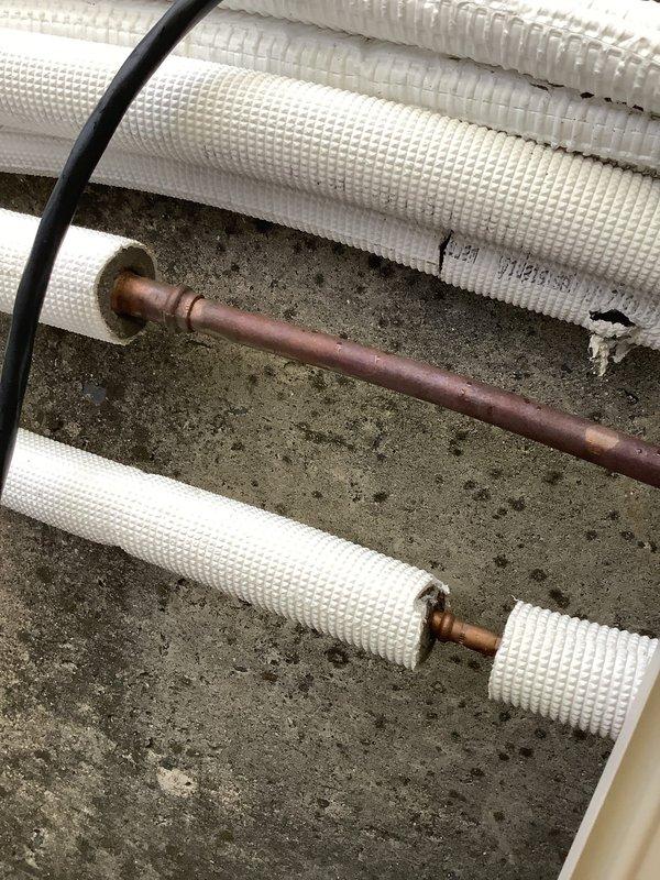 Responded to service call for rooftop HVAC units at commercial property with both office and store units not cooling properly. Used extension ladder to access rooftop equipment located at rear of building. Upon inspection, found units running but not producing cold air. Diagnosed refrigerant system issues evidenced by severely corroded copper refrigerant lines with oxidation and brownish patina indicating age and potential leak points. White foam insulation on refrigerant lines showed multiple gaps and deterioration, exposing compromised copper piping. Inspected Daikin thermostat control panel and noted improper installation with damaged drywall and rough mounting edges that could affect system operation. Documented conditions with photos showing extent of refrigerant line corrosion and thermostat installation deficiencies for repair recommendations.