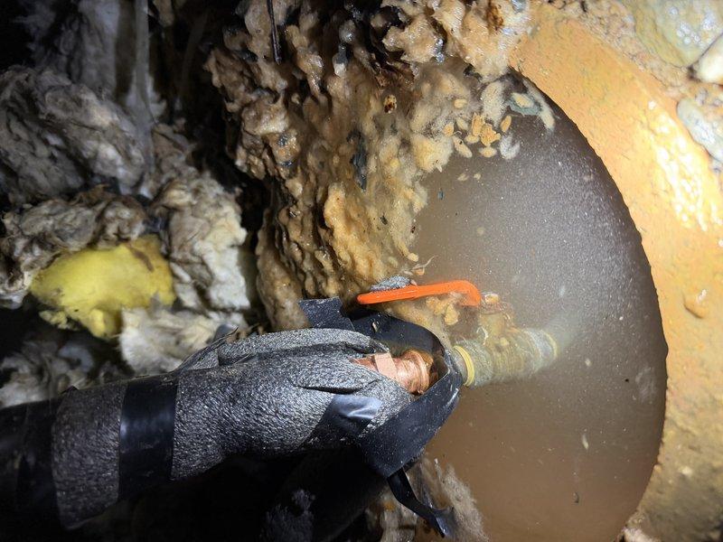 Responded to after-hours emergency call for leaking pipe in crawl space. Upon arrival, accessed utility area and performed electrical testing on wiring near affected plumbing components to ensure safe working conditions. Located source of leak at underground pipe connection point with significant water accumulation and sediment buildup in access pit. Inspected shutoff valve assembly with red handle mechanism and surrounding copper piping. Documented deteriorated conditions including compromised insulation, moisture damage, and debris accumulation throughout work area. Provided photographic documentation of electrical testing procedures and valve access point conditions for estimate preparation and repair planning.