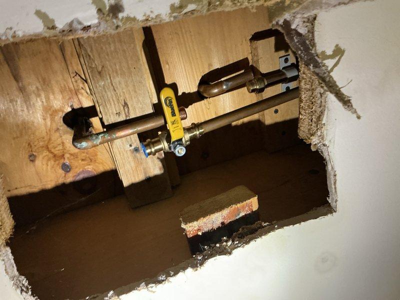 Responded to second-floor bathroom tub leak affecting basement area. Isolated water supply and shut off main line to contain active leak. Cut access opening in closet wall to expose plumbing assembly behind bathtub. Located leak source at Moen tub valve assembly with brass compression fittings and copper supply lines. Previous cartridge replacement had been performed on this fixture. Installed shut-off valve with yellow handle at supply line for isolation purposes. Documented plumbing configuration and framing structure. Coordinated return visit with two-person crew for half-day repair completion to replace faulty components and restore wall opening. Property has secondary shower available for use during repair period.