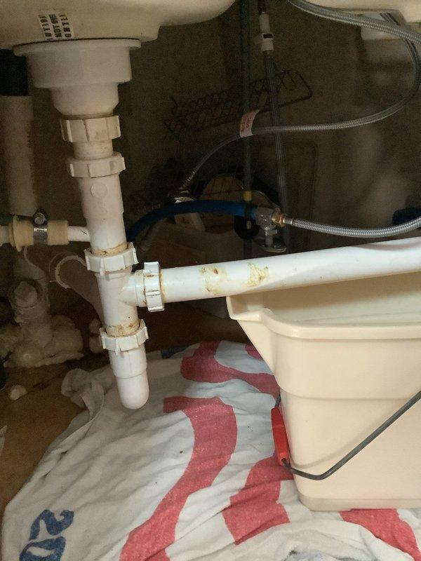 Responded to kitchen sink drain issue with slow drainage and suspected leak beneath fixture. Upon inspection, identified compromised drainage system with visible corrosion staining on PVC piping components. Cleared obstruction from drain line to restore proper flow rate. Inspected under-sink plumbing assembly including basket strainer, P-trap, and horizontal drain connections. Addressed minor leak at pipe joint by tightening connections and replacing worn gasket. Verified water supply lines and flexible braided connections for integrity. Assessed cabinet space for garbage disposal installation compatibility and provided recommendation for future upgrade option.