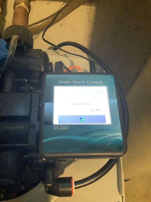 Responded to customer complaint of squealing noise during regeneration cycle, sulfur odor in water, and slimy shower water. Inspected water softener system equipped with Smart Touch Control ST-200 during backwash cycle operation. Verified system regeneration schedule set for 10:30-11:00 PM as configured for household needs. Documented control valve assembly with barcode BND0355940, examined fiberglass tank condition, and checked plumbing connections. Photographed system components including control head, tank assembly, and PVC piping for service records.