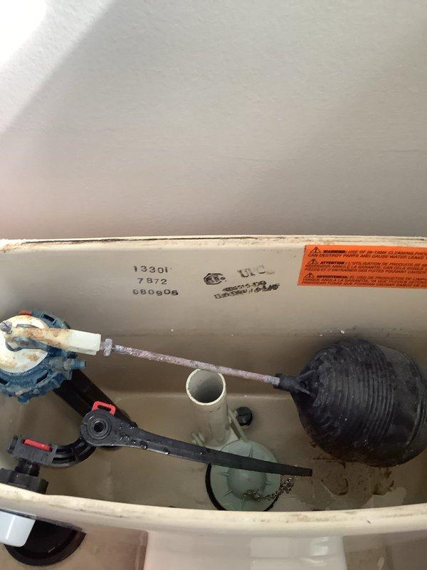Inspected the upstairs toilet for a leak at the base and examined the tank internals, finding a heavily corroded float arm and fill valve with mineral buildup and sediment throughout the tank. Evaluated the supply line and shutoff valve connection and the base seal as potential leak sources, and documented the aged, rusted components as contributing to the water issue.