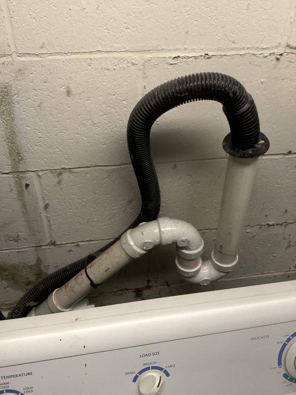 Inspected the washing machine drain connection after a report of water on the floor behind the unit. Found the corrugated drain hose seated in the top of the vertical PVC standpipe with a rubber collar, connected to the P-trap assembly, with staining and debris buildup visible around the lower pipe fittings and the wall. Treated the drain line with acid to clear buildup and restore proper drainage flow, which addresses the likely cause of the standpipe overflow behind the machine.