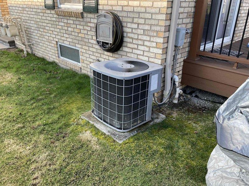 Conducted complimentary estimate for complete HVAC system replacement at residential property. Assessed existing outdoor air conditioning condenser unit with gray casing and black metal grid covering, mounted on concrete pad adjacent to building exterior with refrigerant lines and electrical conduit running along cream-colored brick wall. Evaluated basement heating system and existing infrastructure, noting exposed floor joists, ductwork routing, and current mechanical system layout. Provided comprehensive pricing options for new air conditioning unit and furnace replacement to meet homeowner's comfort needs. Documented existing equipment conditions and installation requirements through photographic records of outdoor condenser unit positioning and basement mechanical space configuration.