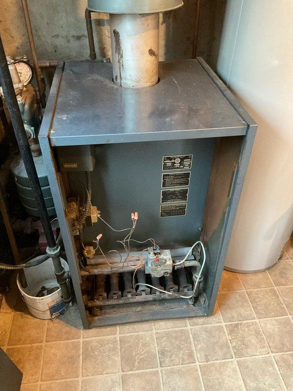 Conducted on-site assessment for boiler replacement in basement utility area. Evaluated existing gray-blue painted boiler unit showing significant age-related deterioration, including dust accumulation on internal components, surface discoloration, and oxidation on associated cylindrical tank. Documented current system configuration including gas valve assembly, copper tubing connections, electrical wiring with wire connectors, and metallic flue pipe extending from unit top. Photographed open access panel revealing internal mechanical components and heating elements for replacement specifications. Prepared comprehensive replacement quote for complete boiler system upgrade with customer requesting expedited scheduling for morning installation window.