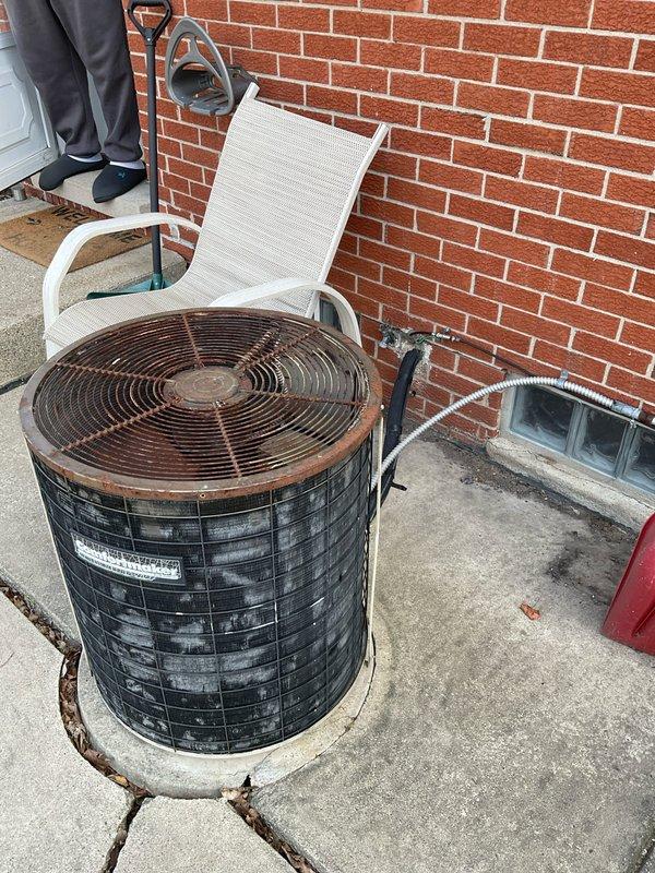 Completed site assessment for HVAC system replacement estimate. Evaluated existing outdoor Goodman condenser unit showing significant rust deterioration on fan grille and housing, with unit positioned on cracked concrete pad against exterior brick wall. Documented indoor Goodman furnace installation in basement utility area with existing ductwork configuration, including metallic supply plenum and insulated return connections. Photographed both components to provide comprehensive system evaluation for replacement quote preparation.