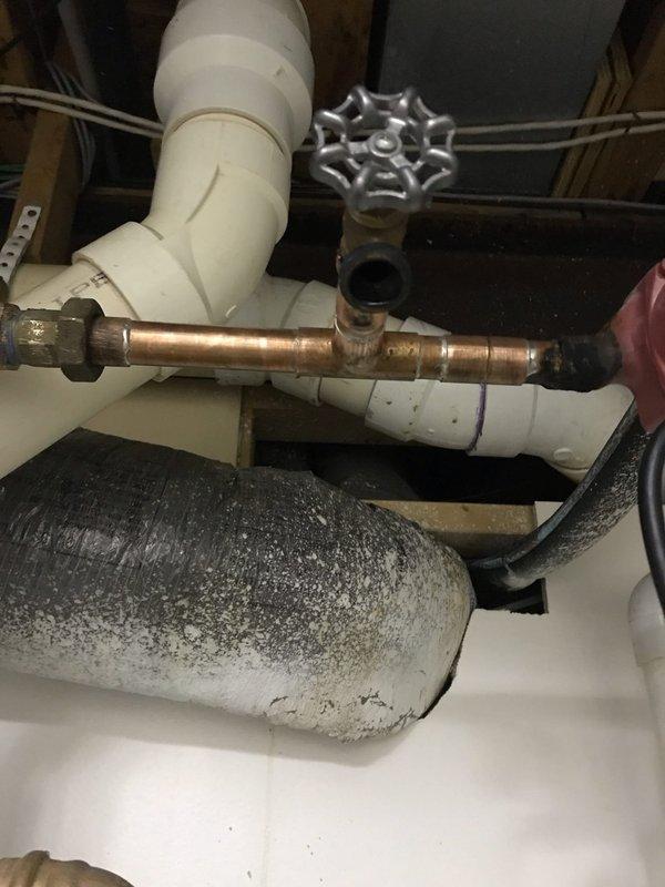 Responded to water heater leak complaint and performed thorough inspection of tanked water heater system and associated plumbing connections. Upon examination, identified source of leak at copper pipe connection point near water heater assembly. Inspected PVC and copper piping configuration, including valve assembly and pump components in utility space. Documented condition of water heater showing surface discoloration and staining consistent with moisture exposure and age-related wear. Photographed plumbing assembly including white PVC elbows, copper piping with chrome valve featuring star-shaped handle, and connection points where leak was occurring. Assessed overall system integrity and identified necessary repairs to address pinhole leak in copper pipe section to prevent further water damage and restore proper system function.