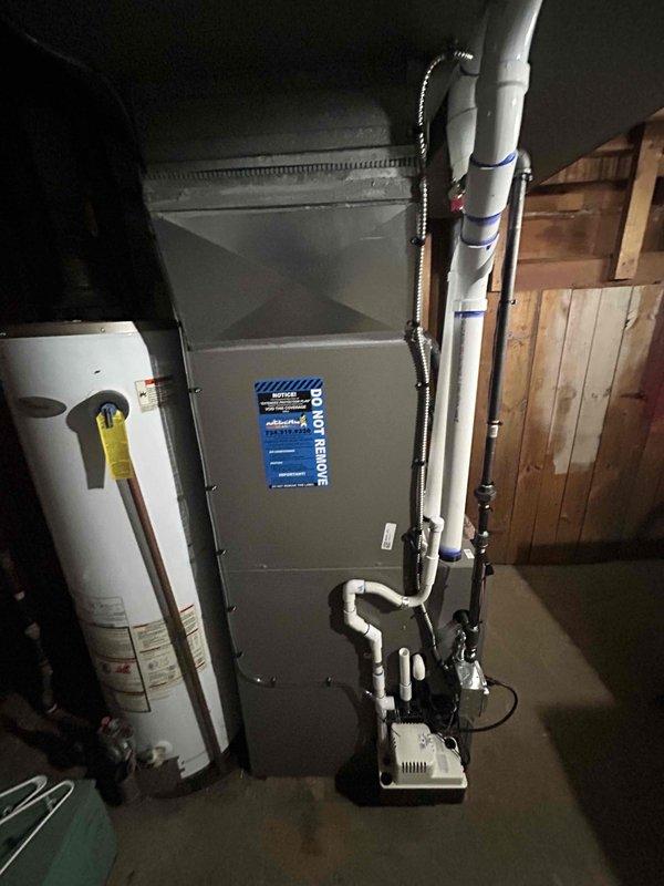 # HVAC System Installation

Successfully completed full HVAC system installation in residential basement utility area. Installed new gray metal furnace unit with proper manufacturer specifications and blue-labeled front panel assembly. Mounted unit on concrete floor with appropriate clearances from surrounding walls and adjacent water heater.

Installed complete white PVC venting and drainage system along right side of furnace, securing all vertical runs with multiple blue straps for proper support and code compliance. Configured PVC condensate drainage piping at base of unit with proper trap assembly to ensure efficient moisture removal. Installed white condensate pump near floor level to facilitate proper drainage operation.

Connected flexible black corrugated supply duct to upper portion of furnace for air distribution. Verified all ductwork connections are secure and properly sealed. Ensured adequate clearance maintained between HVAC equipment and existing water heater with yellow