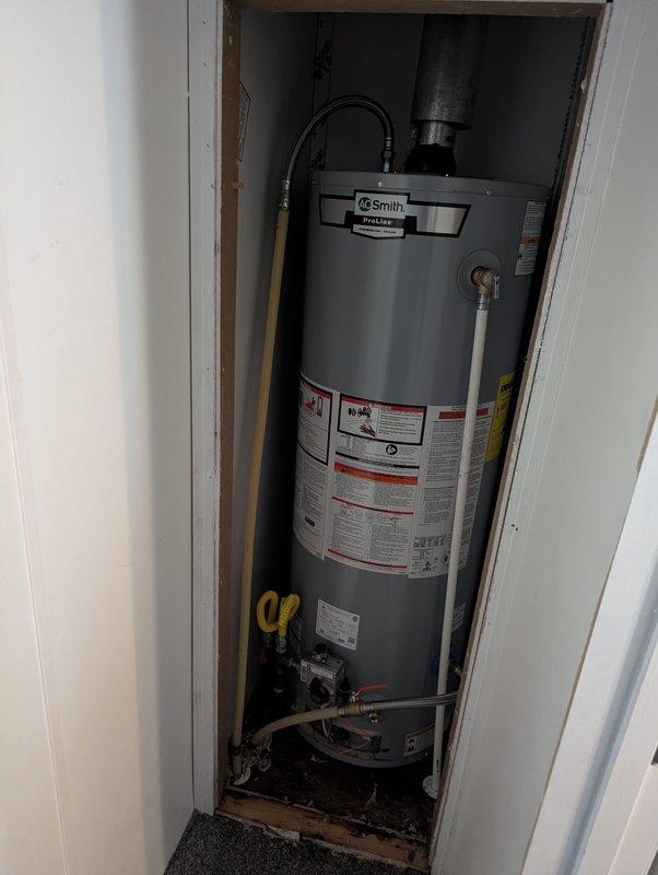 Completed installation of A.O. Smith ProLine residential water heater in confined closet space. Unit properly positioned and secured with all plumbing connections made using copper piping and shut-off valves. Black flexible venting installed and connected to exhaust system at top of unit. All safety labels and specifications verified visible and intact. Base connections completed with yellow-handled isolation valves for future service access.