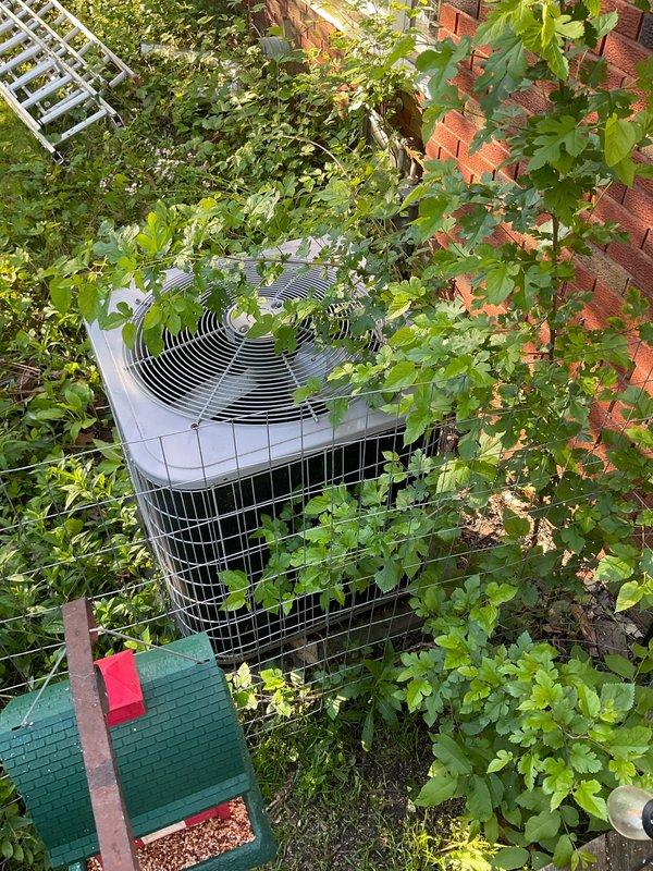 Conducted on-site assessment of existing outdoor air conditioning condenser unit for replacement estimate. Unit located in residential backyard setting with significant vegetation overgrowth impeding proper airflow and accessibility. Documented current equipment condition showing cylindrical cage-style condenser with compromised clearance due to dense vines and plants growing around and over the unit. Evaluated installation site constraints including proximity to brick building foundation, adjacent white lattice fencing, and garden furniture placement. Captured photographic documentation of unit positioning, surrounding obstructions, and access points for potential same-day replacement installation. Provided comprehensive system replacement quote addressing necessary vegetation clearance and proper equipment spacing requirements per manufacturer specifications.