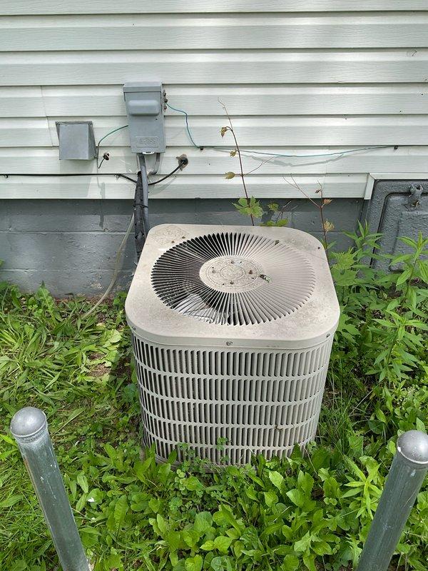 Conducted on-site assessment of existing outdoor air conditioning condenser unit for replacement estimate. Documented current system configuration including beige/tan condenser with spiral fan grille positioned against exterior wall with electrical disconnect boxes and wiring. Evaluated unit accessibility and noted vegetation overgrowth around equipment base requiring clearance for proper airflow and service access. Provided comprehensive estimate options for single component AC replacement and complete system replacement to address customer's cooling needs.