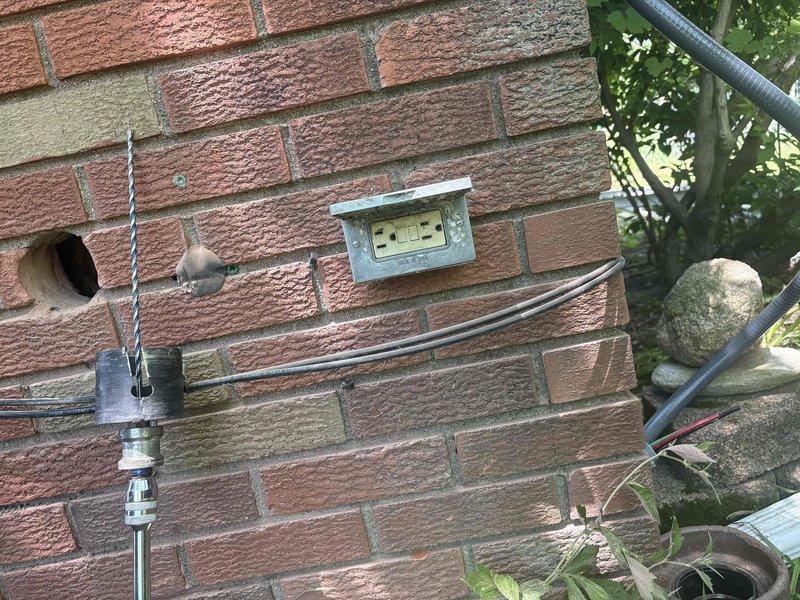 Installed high voltage electrical line from main service panel to exterior outlet location. Ran new electrical conduit through basement utility area, routing orange-jacketed high voltage cable from breaker panel through cinder block wall penetration to outdoor mounting point on brick exterior. Added new circuit breaker to existing electrical panel and terminated connections per code requirements. Installed exterior weatherproof outlet box on brick facade with proper conduit protection and secure mounting hardware. Verified all connections, tested circuit functionality, and ensured proper grounding throughout the installation.