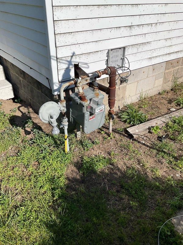 Conducted comprehensive site assessment for HVAC system replacement at residential property. Evaluated existing outdoor utility installation including water heater, associated piping infrastructure consisting of black iron, copper, and PVC materials, and electrical control components mounted on exterior siding. Documented current equipment configuration, pipe routing, and connection points for replacement planning. Assessed structural conditions including foundation access, utility placement on ground level, and clearance requirements for new equipment installation. Photographed existing system layout and surrounding lawn area with visible PVC conduit penetration to establish baseline conditions and facilitate accurate equipment sizing and replacement specifications for realtor transaction.