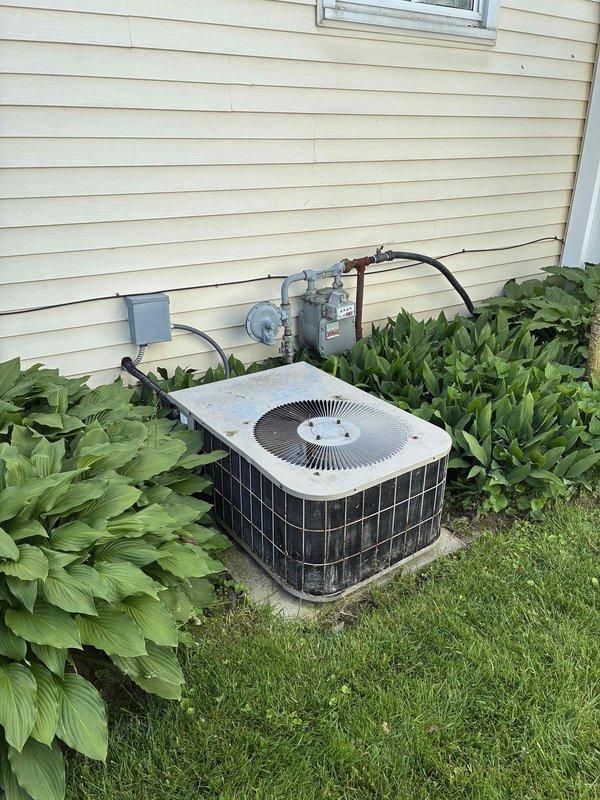 Conducted comprehensive site evaluation for complete HVAC system replacement at residential property. Documented existing outdoor condensing unit with standard configuration including white top cover, circular fan grille, and protective black metal side panels. Unit currently positioned on grade-level installation with direct wall mounting and electrical disconnect box present. Assessed electrical service panel containing dual-row circuit breaker configuration with labeled circuits for proper system integration planning. Evaluated existing refrigerant line set routing, condensate drainage pathway, and electrical conduit runs from exterior unit to main panel. Inspected clearance requirements around outdoor equipment, noting adequate space from landscaping and building structure for proper airflow and service access. Prepared detailed estimate documentation for 10-12 ton system replacement including equipment specifications, installation requirements, and necessary electrical modificatio