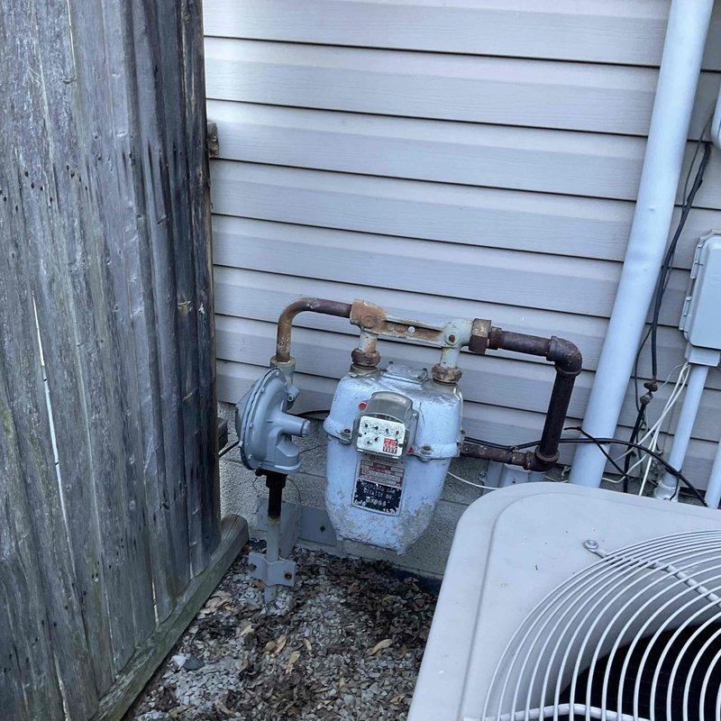 Conducted on-site evaluation for HVAC system replacement at residential property. Documented existing outdoor condenser unit location adjacent to gas meter and electrical service panel on side of building with vinyl siding. Photographed current utility configuration including weathered wooden fence line, gravel pad area, and existing electrical meter assembly with conduit routing to assess installation requirements and clearances for proposed equipment replacement.