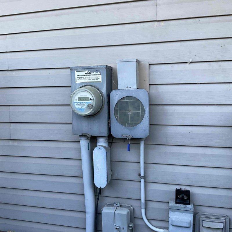 Conducted on-site evaluation for HVAC system replacement at residential property. Documented existing outdoor condenser unit location adjacent to gas meter and electrical service panel on side of building with vinyl siding. Photographed current utility configuration including weathered wooden fence line, gravel pad area, and existing electrical meter assembly with conduit routing to assess installation requirements and clearances for proposed equipment replacement.