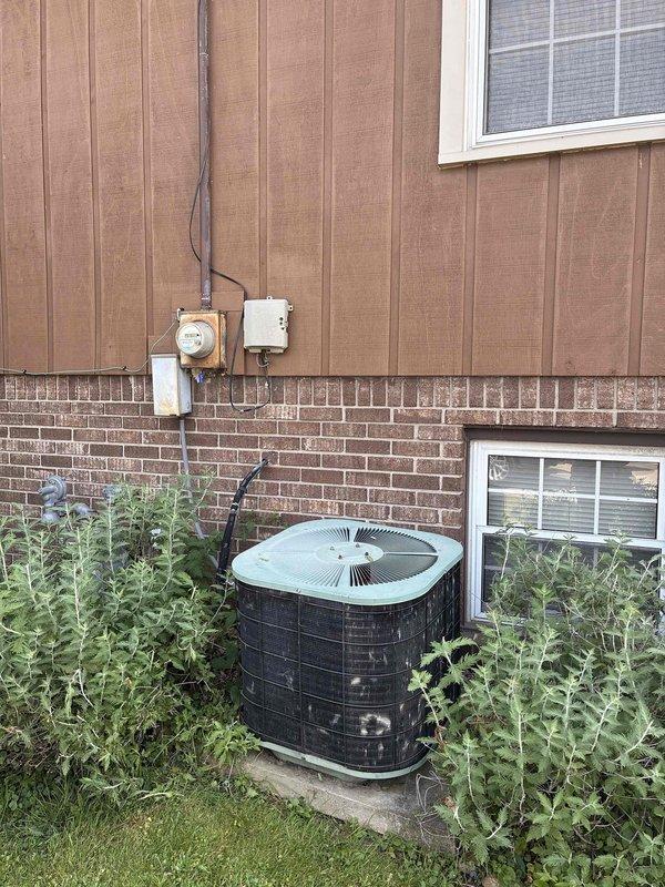Provided estimate for complete air conditioning system replacement on 25-year-old non-operational unit located at exterior of residence. Assessed existing outdoor condenser unit mounted on concrete pad with overgrown vegetation requiring clearance, evaluated electrical service components including meter, disconnect, and conduit routing along brown siding and brick exterior wall. Quoted replacement system compatible with existing natural gas furnace infrastructure.